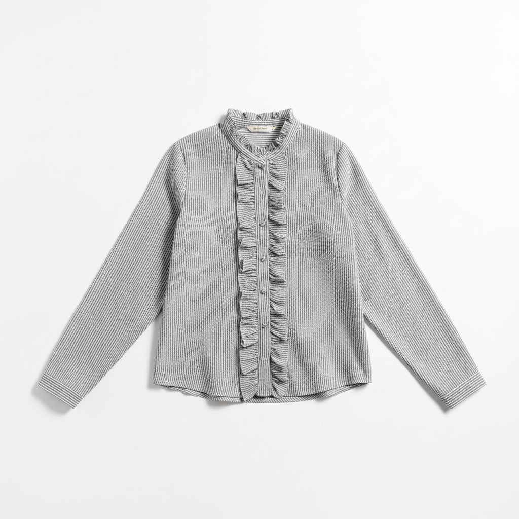 A woman wearing a long-sleeve button-front blouse with vertical ruffle detailing and a gray-and-white stripe pattern, paired with light blue jeans in a bright living room setting.
