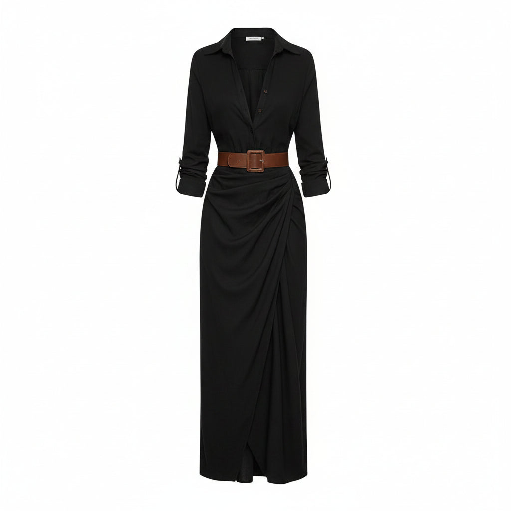 A woman models a long black button-front shirt dress with a deep V-neck, long sleeves, and a matching long skirt. She wears a brown belt with a large square buckle at the waist and carries a woven straw tote bag against a textured light gray wall.